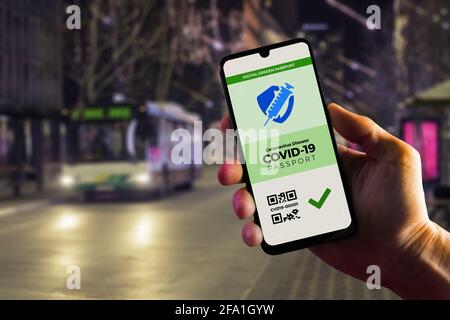 Smartphone zeigt einen gültigen digitalen grünen Pass für COVID-19 in männlicher Hand, Innenstadt und Stadtbus im Hintergrund. Impfung, Krankheitsimmunität p Stockfoto