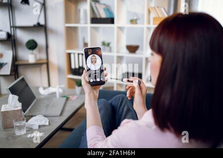 Telemedizinisches Konzept. Rückansicht einer kranken kaukasischen Frau, die zu Hause auf dem Sofa sitzt und ihre Ärztin anruft, während sie das Thermometer in der Hand hält. Tisch mit Laptop und Medikamenten auf dem Hintergrund. Stockfoto
