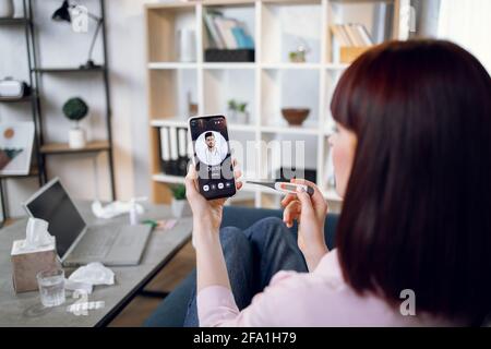 Konzept der Telemedizin und Patientenberatung online. Kranke Frau, die sich zu Hause schlecht fühlt, die Körpertemperatur misst, mit Smartphon zum indischen Arzt ruft. Konzentrieren Sie sich auf den Telefonbildschirm Stockfoto