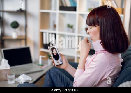 Junge kranke Frau, die Halsschmerzen, Schmerzen und Husten hat, zu Hause auf der Couch sitzt und eine Videobefragung mit einem Arzt, einer arabischen Dame im Hijab, hat. Online-Arzttermin. Stockfoto