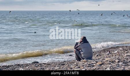 Ein älterer Mann in einer grauen Jacke und einem Strickhut sitzt einsam auf einem felsigen Strand in der Nähe des kalten Meeres und blickt auf das Wasser und die Möwen. Stockfoto