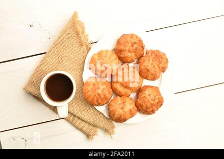 Mehrere frisch duftende Chouquettes auf einer weißen Untertasse mit einer Tasse Kaffee, Nahaufnahme, auf einem weißen Holztisch, Draufsicht. Stockfoto