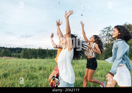 Eine Gruppe multiethnischer Freundinnen hob die Hände hoch und warf sie Konfetti beim Laufen auf einem Feld Stockfoto