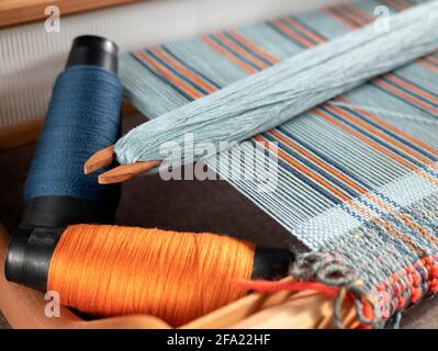 Handloom mit Shuttle auf den blauen Kettfäden und zwei Spulen mit Indigo- und Orange-Garnen. Vertikales Streifengewebe Stockfoto