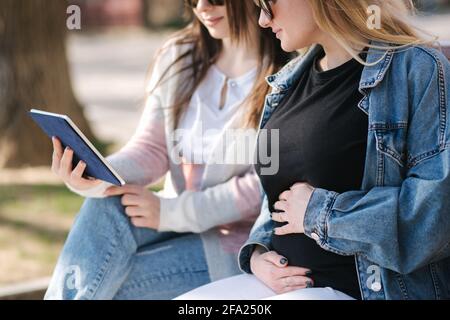 Eine Schwangere mit ihrem besten Freund, der im Park sitzt und mit einem Tablet nach einem Kinderwagen sucht. Zwei Frauen in Sonnenbrillen wählen Kleidung für das Baby Stockfoto