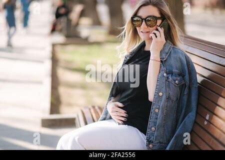 Die Frau von Pregnat sitzt auf der Bank im Park und telefoniert. Glückliche Weibchen haben Ruhe draußen während der Spaziergänge Stockfoto
