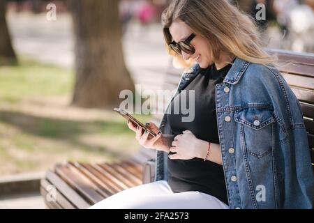 Die Frau von Pregnat sitzt auf der Bank im Park und telefoniert. Glückliche Weibchen haben Ruhe draußen während der Spaziergänge Stockfoto