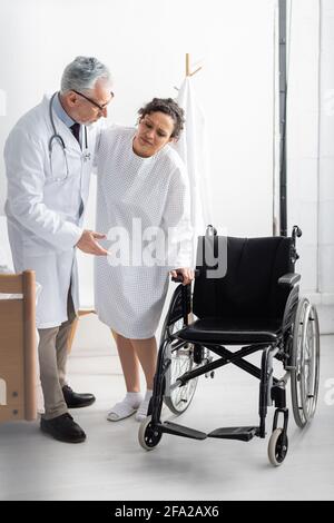 Doktor zeigt auf Rollstuhl, während er afroamerikanische Frau unterstützt Krankenhaus Stockfoto