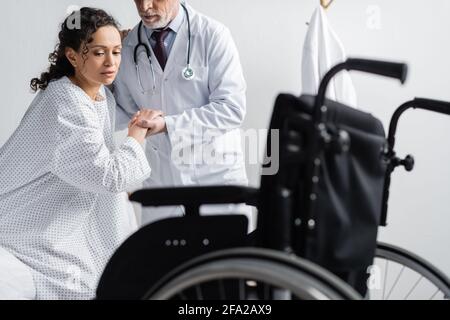 Arzt, der afroamerikanische Frau in der Nähe des Rollstuhls auf verschwommenem Vordergrund unterstützt Stockfoto