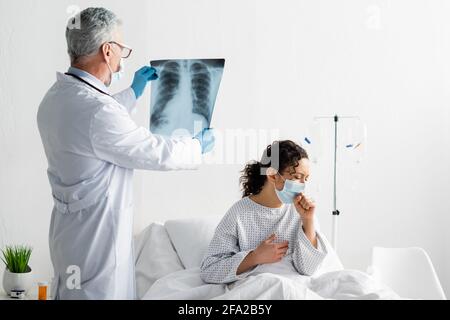 Radiologe in der medizinischen Maske hält Lunge Röntgen in der Nähe von afroamerikaner Maskenhusten auf Krankenhausbett Stockfoto