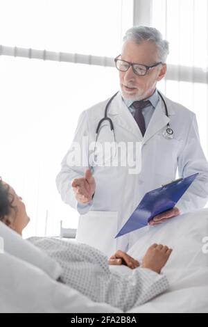 Reifer Arzt zeigt mit der Hand, während er mit afroamerikaner spricht Frau auf verschwommenem Vordergrund Stockfoto