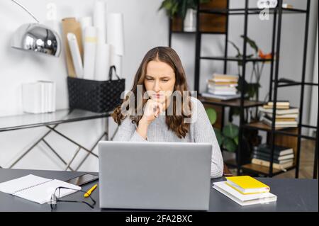 Konzentrierte Frau starrt auf den Laptop-Bildschirm, der im Büro sitzt, eine nachdenkliche, fokussierte Mitarbeiterin, die nach der Lösung von Projektaufgaben sucht und die per E-Mail erhaltenen Einverständungsbedingungen sorgfältig liest Stockfoto