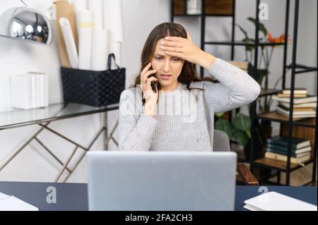 Besorgte junge Geschäftsfrau, die verzweifelt den Kopf hält, auf den Laptop-Bildschirm schaut und am Telefon telefoniert, müde und stressig Stockfoto