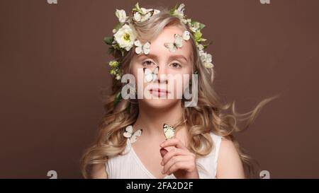 Hübsche junge Frau mit gesunden blonden, windigen Haaren, weißen, sanften Blumen und Schmetterling in der Hand auf braunem Hintergrund Stockfoto