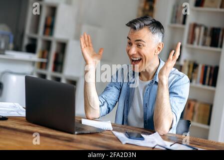 Überglücklich, grauhaariger kaukasischer Freiberufler, Manager, in legerer Kleidung gekleidet, mit Laptop, freut sich über Erfolg, Sieg oder guten Gewinn, schaut auf einen Laptop-Bildschirm und Gesten mit seinen Händen Stockfoto