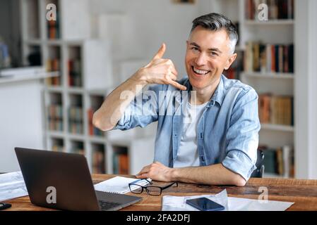 Freundliche, gesellige, selbstbewusste, grauhaarige kaukasische Freiberuflerin mittleren Alters, in stilvoller Kleidung, arbeitet fernab an einem Laptop, zeigt die „Call Me“-Geste, blickt auf die Kamera und lächelt Stockfoto