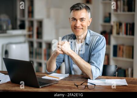 Porträt eines selbstbewussten, intelligenten grauhaarigen, reifen kaukasischen Freiberuflers, Maklers oder Managers, in stilvoller Freizeitkleidung, der zu Hause auf einem Laptop arbeitet, am Schreibtisch sitzt, die Kamera anschaut und freundlich lächelt Stockfoto