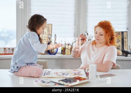 Glückliche Familie zusammen: Mutter und Tochter malen zu Hause mit Pinsel und Aquarell zusammen. Junge Frau sitzt und hilft oder lehrt Tochter auf einem Tisch sitzen zu zeichnen Stockfoto