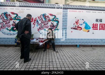 Eine ältere Frau plaudert mit einem Mann, während sie hinter ihr ein Kind von Propagandaplakaten auf einer Wand sitzt, Einlagen für Schuhe verkauft und Textilien in den Straßen von Urumqi in Xinjiang, China, VR China, fixiert. © Time-Snaps Stockfoto