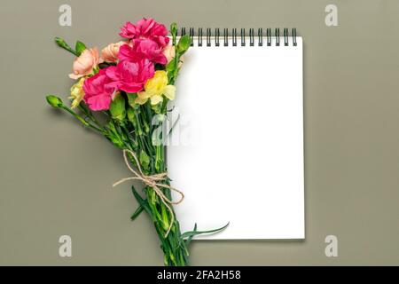 Farbenfroher Strauß aus verschiedenen rosa Nelkenblumen, weißes Notizbuch auf grünem Hintergrund Draufsicht Flat Lay Weihnachtskarte 8. März, Happy Valentine's day Stockfoto