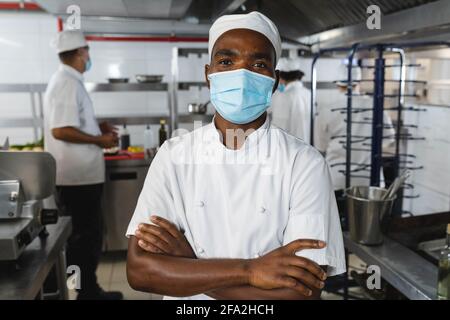 Porträt eines afroamerikanischen professionellen Küchenchefs mit Gesichtsmaske Mit Kollegen im Hintergrund Stockfoto