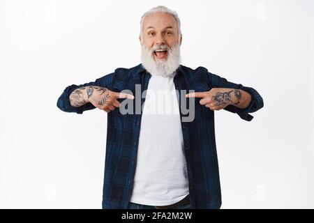 Ein alter glücklicher Mann mit Tattoos und langem grauen Bart, der mit aufgeregt lächelndem Gesicht auf sich zeigt, ein Tor gewinnt oder erreicht, hört von guten Neuigkeiten Stockfoto