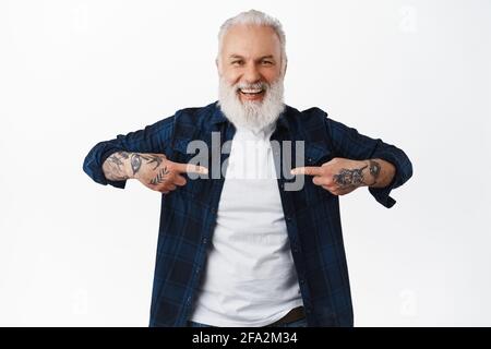 Schöner älterer Mann, alter Mann, der auf sich zeigt und lacht. Großvater mit Tattoos und langem Bart selbstfördernd, prahlend oder prahlend, stehend Stockfoto