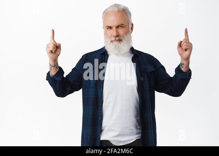 Schöner alter Mann mit Tattoos und Bart, zeigt den Finger nach oben und lächelt, gibt Ratschläge, um ein Produkt zu kaufen, zeigt Werbung oder Logo Ihrer Marke Stockfoto