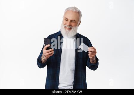 Schöner reifer Mann, der mit Kreditkarte auf dem Smartphone bezahlt, Karte zeigt und mit lächelndem Gesicht auf das Telefon schaut, online einkauft, dagegen steht Stockfoto