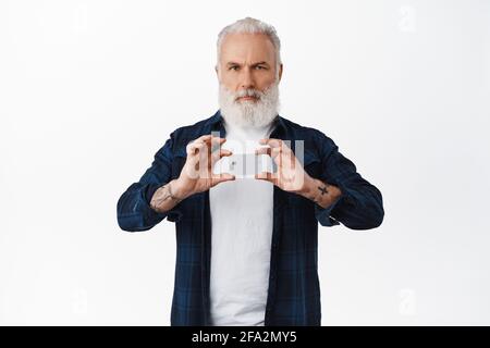 Seriöser älterer Mann mit langem weißen Bart und Tattoos, zeigt Kreditkarte und Stirnrunzeln selbstbewusst, steht vor weißem Hintergrund Stockfoto