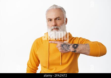 Cooler älterer Hipster-Mann mit Tattoos, die nach links auf den Kopierbereich zeigen, mit Werbung, Großvater mit langem Bart und schickem Hoodie Stockfoto