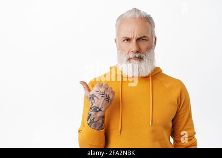 Enttäuschter, ernster alter Mann mit Tattoos, der nach links zeigt und wütend auf die Kamera schaut, Fragen zur Logo-Marke stellt, Erklärungen braucht, schimpft Stockfoto