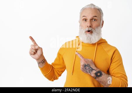 Stilvoller, moderner Seniorchef, der auf die linke obere Ecke zeigt und überrascht und interessiert aussieht. Alter Typ mit Tattoos und langem Bart, der Fragen stellt Stockfoto