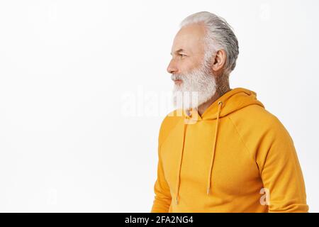 Stilvoller, hübscher Senior mit langem Bart, orangefarbenem Hoodie, drehe den Kopf nach links und schaue auf leeren Raum für dein Logo oder Werbebanner Stockfoto