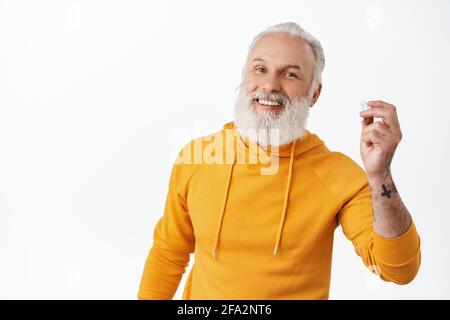 Lächelnder Hipster alter Mann zieht den Kopfhörer ab und schaut glücklich aus, stellt einen Kopfhörer in das Ohr, um Musik zu hören, während man telefonieren kann, und nutzt kabellose Ohrstöpsel Stockfoto