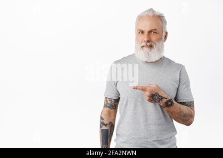 Stilvoller reifer Mann mit Tattoos, die mit dem Finger nach links zeigen. Hipster alter Kerl mit langem Bart, der Werbung zeigt und zufrieden lächelt und Firma empfiehlt Stockfoto