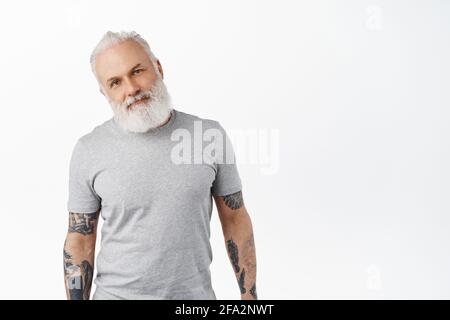 Stilvoller, authentischer Seniorchef mit Tattoos und langem Bart, im lässigen grauen T-Shirt stehend, glücklich und zufrieden in der Kamera, lächelnd, überstehend Stockfoto