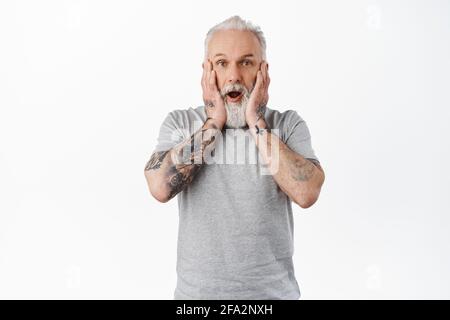 Schockiert reifer Mann mit Tattoos, keuchen und sehen alarmiert aus, halten Hände auf das Gesicht und starren erschrocken und besorgt auf die Kamera, hören schlechte Nachrichten, stehen Stockfoto