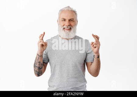 Daumen drücken. Lächelnder älterer Mann mit Tattoos, die Wünsche machen, optimistisch und hoffnungsvoll aussehen, auf gute Nachrichten warten, Träume wahr werden, hereinstehen Stockfoto