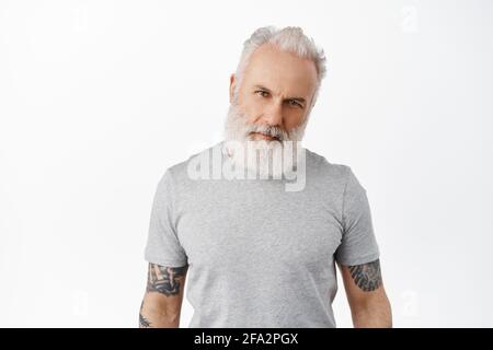 Stilvoller, hübscher, reifer Mann mit tätowiertem Körper und langem Bart, neigtem Kopf und selbstbewusster Macho-Optik auf die Kamera, ernst anstarrend und in grauem T-Shirt stehend Stockfoto