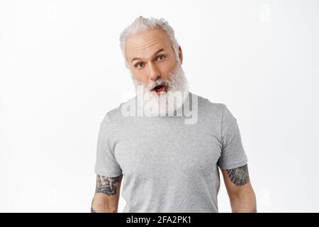 Überraschender, gutaussehender alter Mann lässt den Kiefer fallen, schnappt vor Ehrfurcht, starrt auf etwas Erstaunliches, schaut sich Werbung an, steht in grauem T-Shirt gegen Weiß Stockfoto