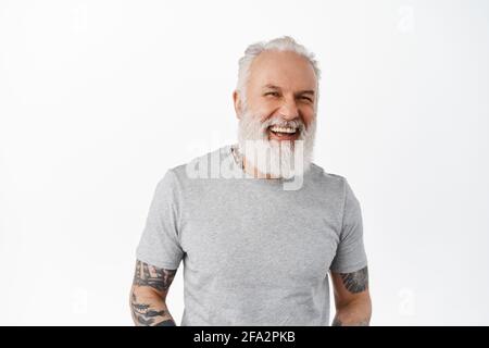 Nahaufnahme eines gutaussehenden älteren Mannes mit Tätowierungen und Bart, lachend und offen zur Seite schauend, plaudernd und lustig, glücklich gegen Weiß stehend Stockfoto