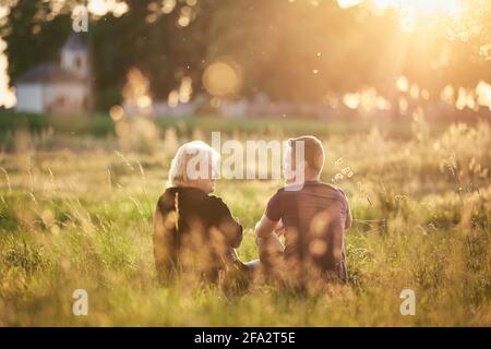 Mutter sitzt mit Sohn auf der Wiese. Ältere Frau und junger erwachsener Mann sprechen bei Sonnenuntergang gegen das Dorf. Stockfoto