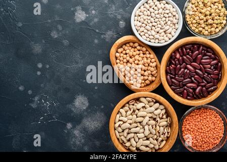 Hülsenfrüchte, rote Linsen, Kichererbsen, rote und weiße Bohnen werden in verschiedenen Schüsseln auf einem dunkelschwarzen Tisch angeboten. Diät zur Gewichtsabnahme und Kampf gegen die Choleste Stockfoto