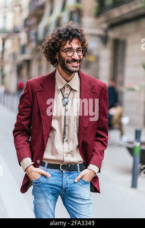 Porträt eines attraktiven kaukasischen jungen Mannes, der in den Straßen von Barcelona mit den Händen in den Taschen steht. Er lächelt und schaut zur Seite. Er hat lockiges Haar, Bart und Brille. Modische Freizeitkleidung Stockfoto