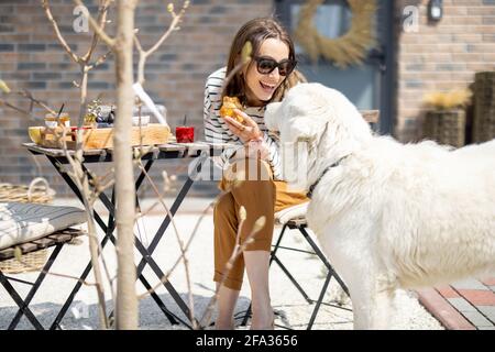 Fröhliche Frau, die mit einem großen weißen Hund an einem Tisch im Garten neben dem Haus sitzt und zu Mittag ass. Verbringen Sie Zeit im Freien. Stockfoto