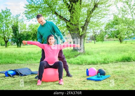 Ein Paar lächelnder persönlicher Trainer und eine schöne Schwangeren, die Workout-Übungen im Freien in einem Park machen.Girl, das Gewicht hebt, sitzt auf dem Gymnastikball. Stockfoto