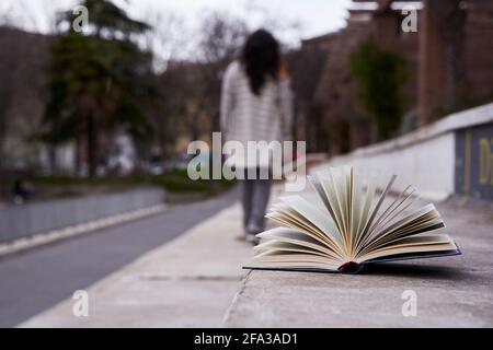 Offenes Buch, vergessen auf einem Steinsitz und eine Frau, die unfokussiert davonging. Stockfoto