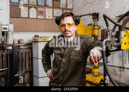 Porträt eines jungen modernen Handwerkers vor dem Hintergrund einer Schmiede, eines Mannes in gelben Schutzbrillen und Kopfhörern Stockfoto