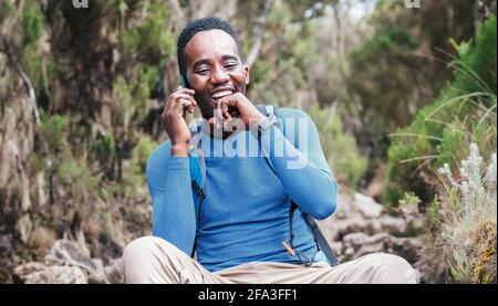 Junger afrikanischer Ethnie Mann, der per Handy mit jemandem redet und fröhlich lachend, als er einen Wanderspaziergang im tropischen Wald macht. Viel Spaß Stockfoto